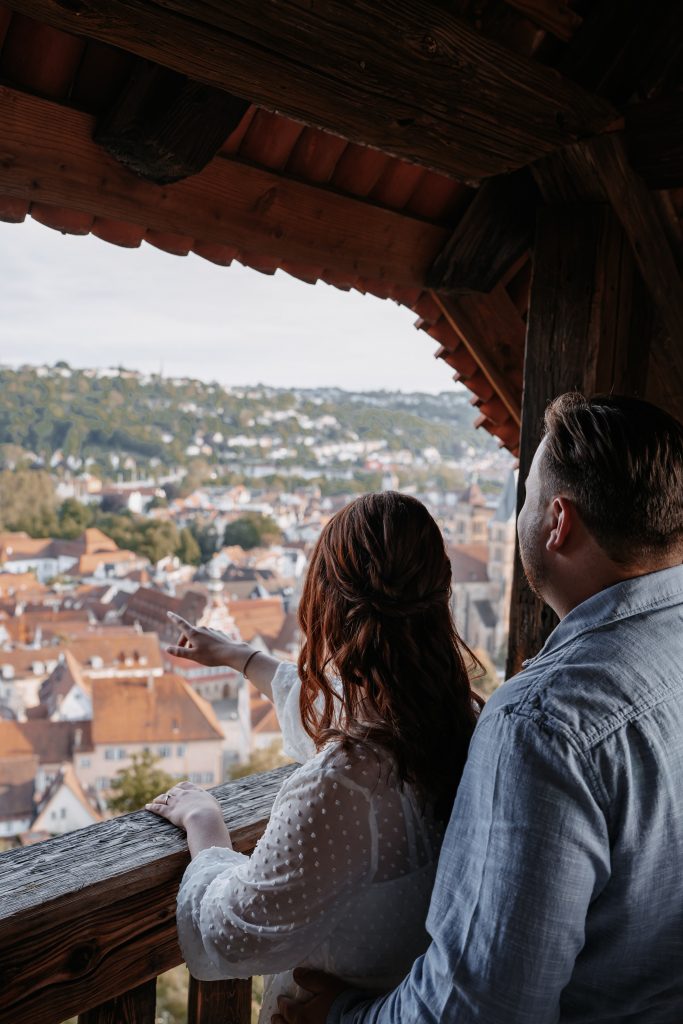Ein Paar steht auf einem hölzernen Aussichtsbalkon und blickt über eine Stadt mit roten Dächern und grüner Hügellandschaft.